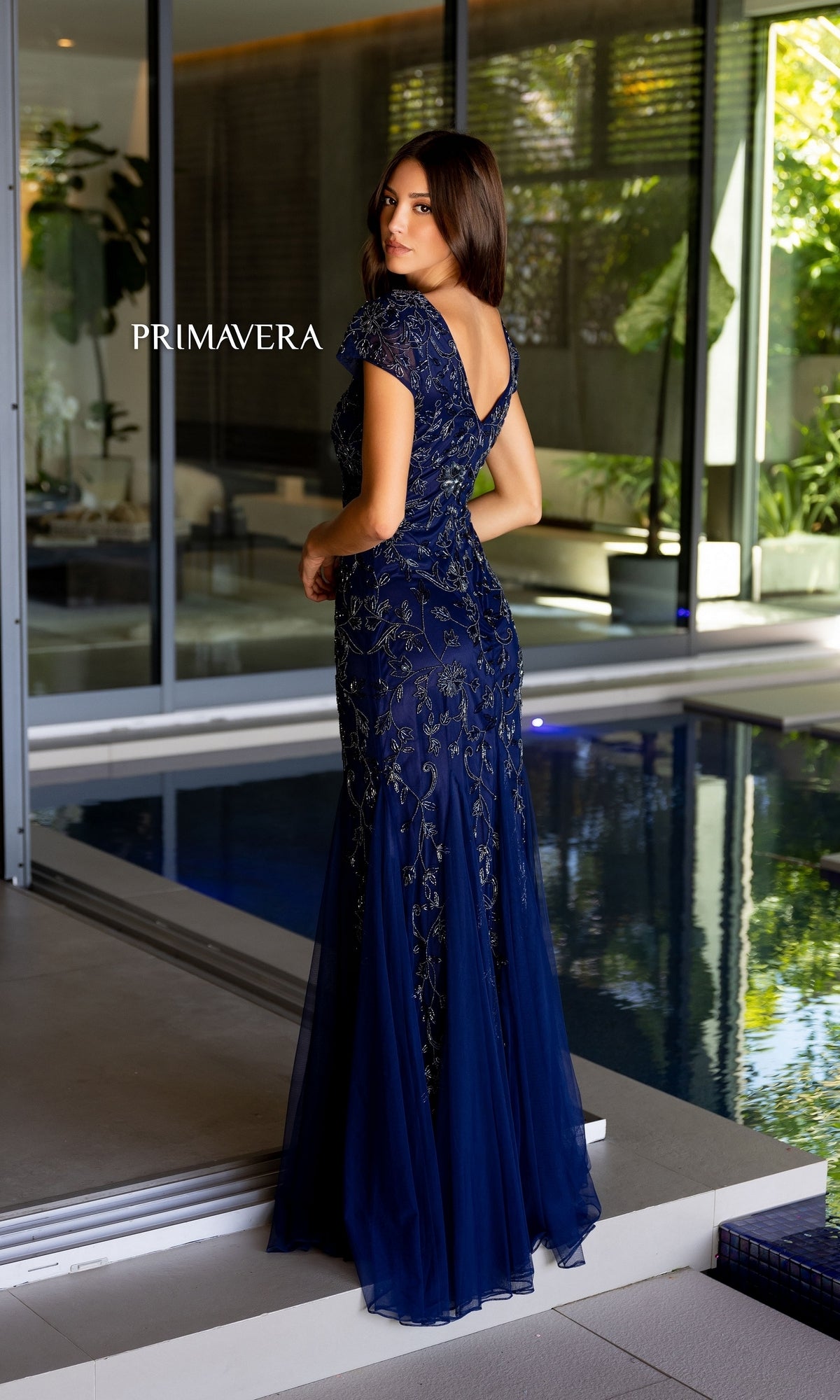 A woman stands near a modern indoor pool, facing away, wearing the Long Prom Dress 13112 by Primavera—a blue, floral beaded mermaid gown.